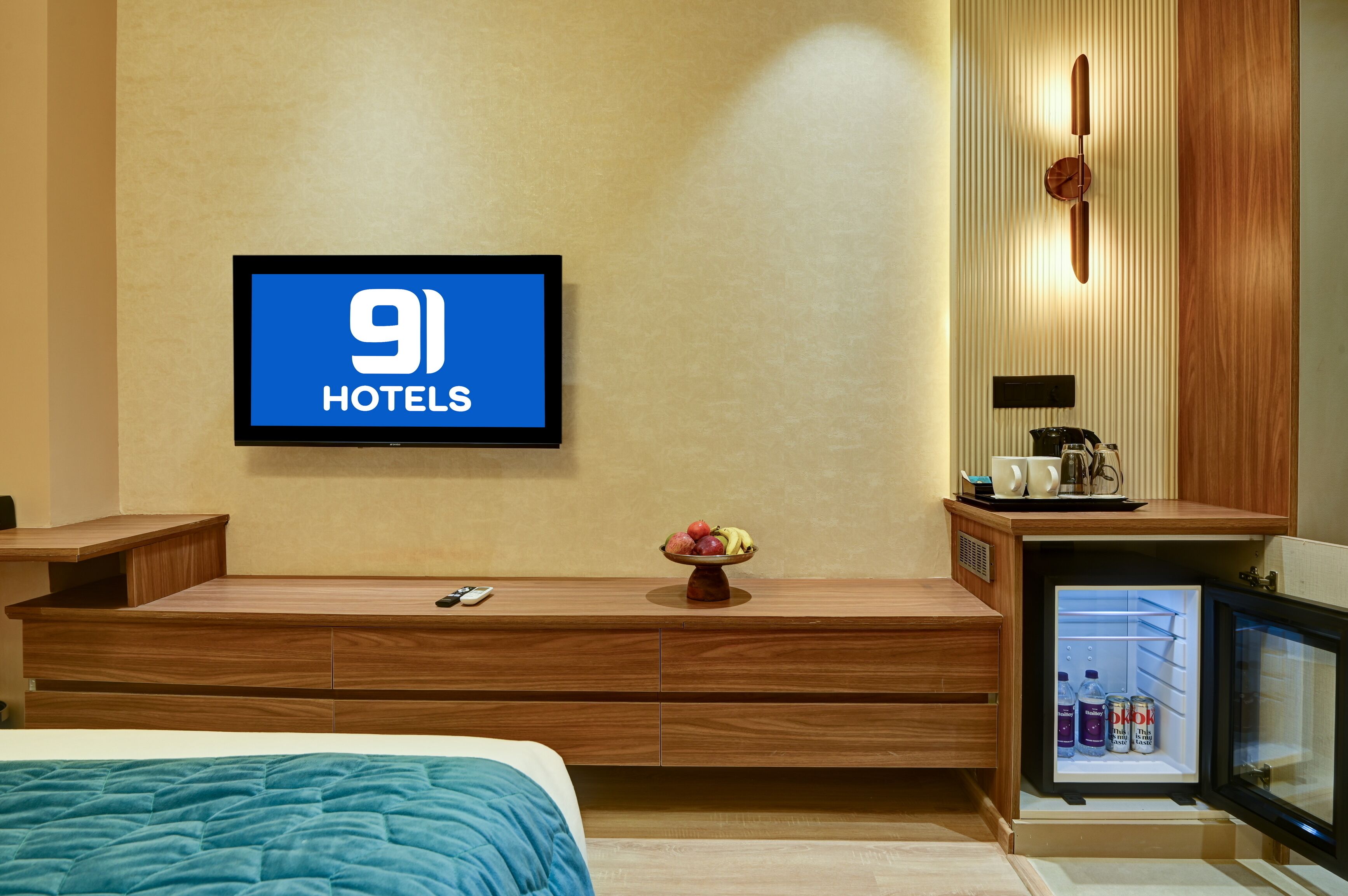 Camera d'albergo con un letto, una TV che mostra '9 Hotels', un tavolino con una ciotola di frutta, una macchina del caffè e un mini-frigo con bevande.