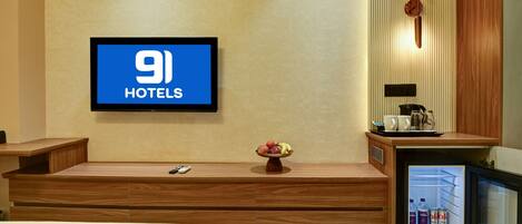 Camera d'albergo con un letto, una TV che mostra '9 Hotels', un tavolino con una ciotola di frutta, una macchina del caffè e un mini-frigo con bevande.