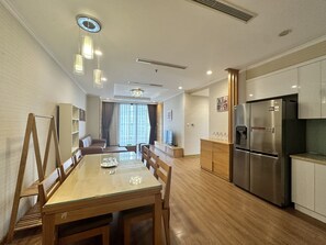A modern kitchen with a stainless steel refrigerator, wooden cabinets, and a dining area with a glass table and chairs. - Vinhomes Royal City by Emily in Town (Hanoi)