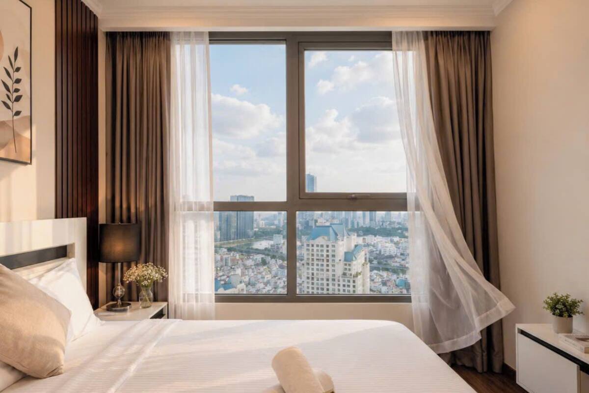 A hotel room with a large bed, a nightstand with a lamp, a view of a cityscape through a large window, and a small table with a plant.