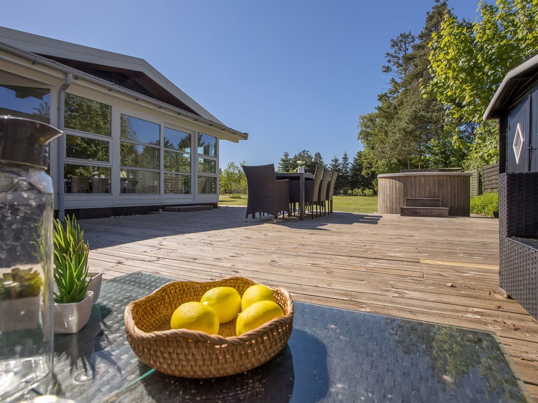 Una terraza de madera con una mesa de vidrio, una cesta de mimbre con limones y mobiliario de exterior.