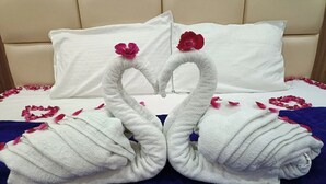 Ein Bett mit weißen Kissen und einer blauen Decke, verziert mit zwei schwanenförmigen Handtüchern und roten Rosenblättern.