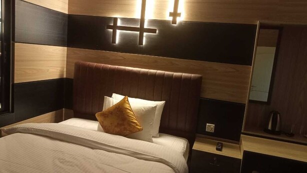 A hotel room with a bed, a nightstand, a mirror, and a cross-shaped light fixture on the wall. - Garden View Villa (Rajshahi)