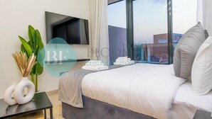 Habitación de hotel moderna con una cama grande, un televisor de pantalla plana, una planta en maceta y vistas de la ciudad a través de amplias ventanas.