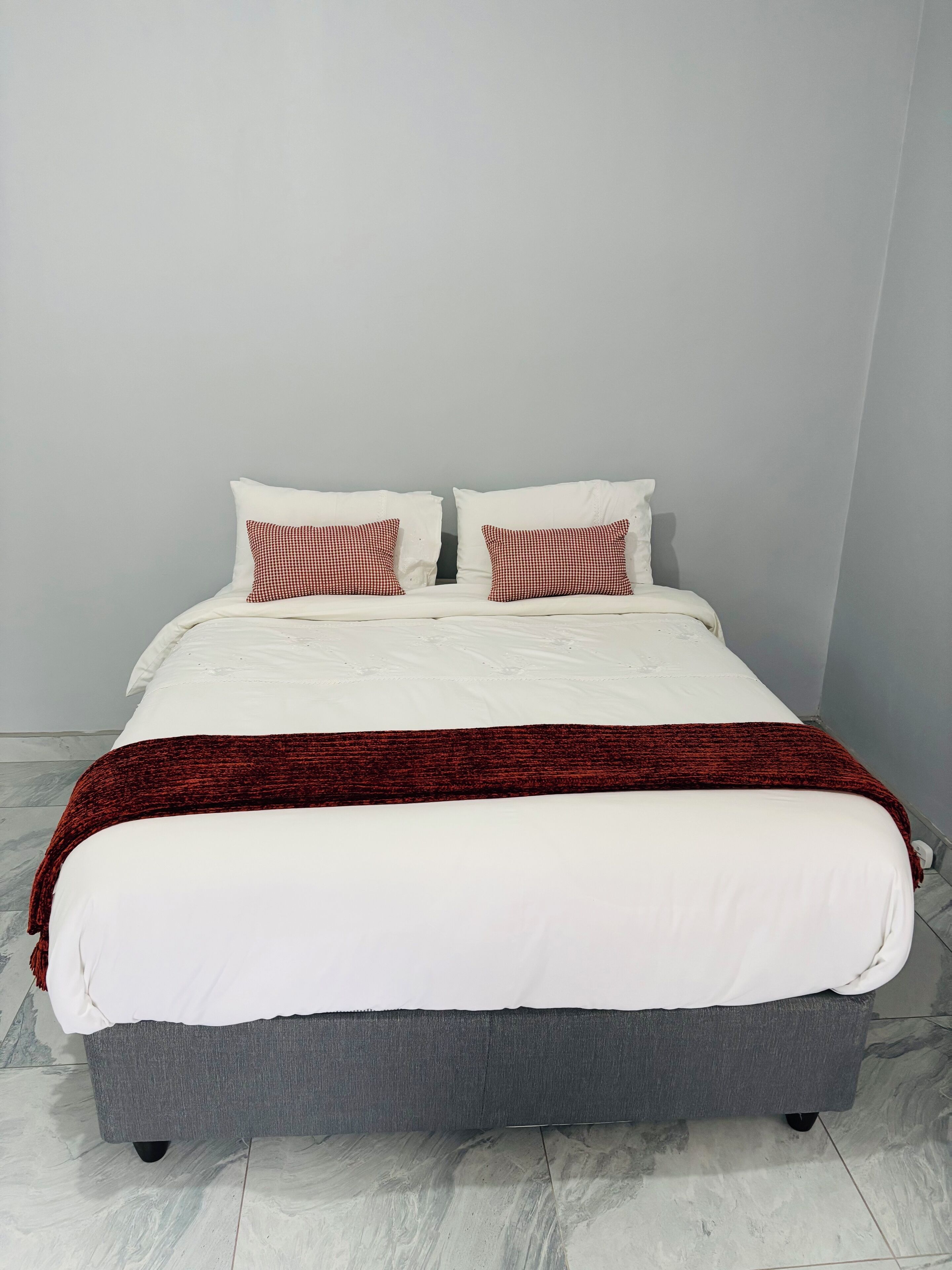 Ein Bett mit weißer Bettwäsche und einer roten Decke, zwei Kissen mit Karomuster sowie ein grauer, gepolsterter Kopfteil.