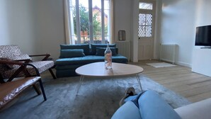 Una sala de estar con sofá, mesa de centro y una silla. Se ve una ventana con cortinas, un radiador y un televisor en la pared.