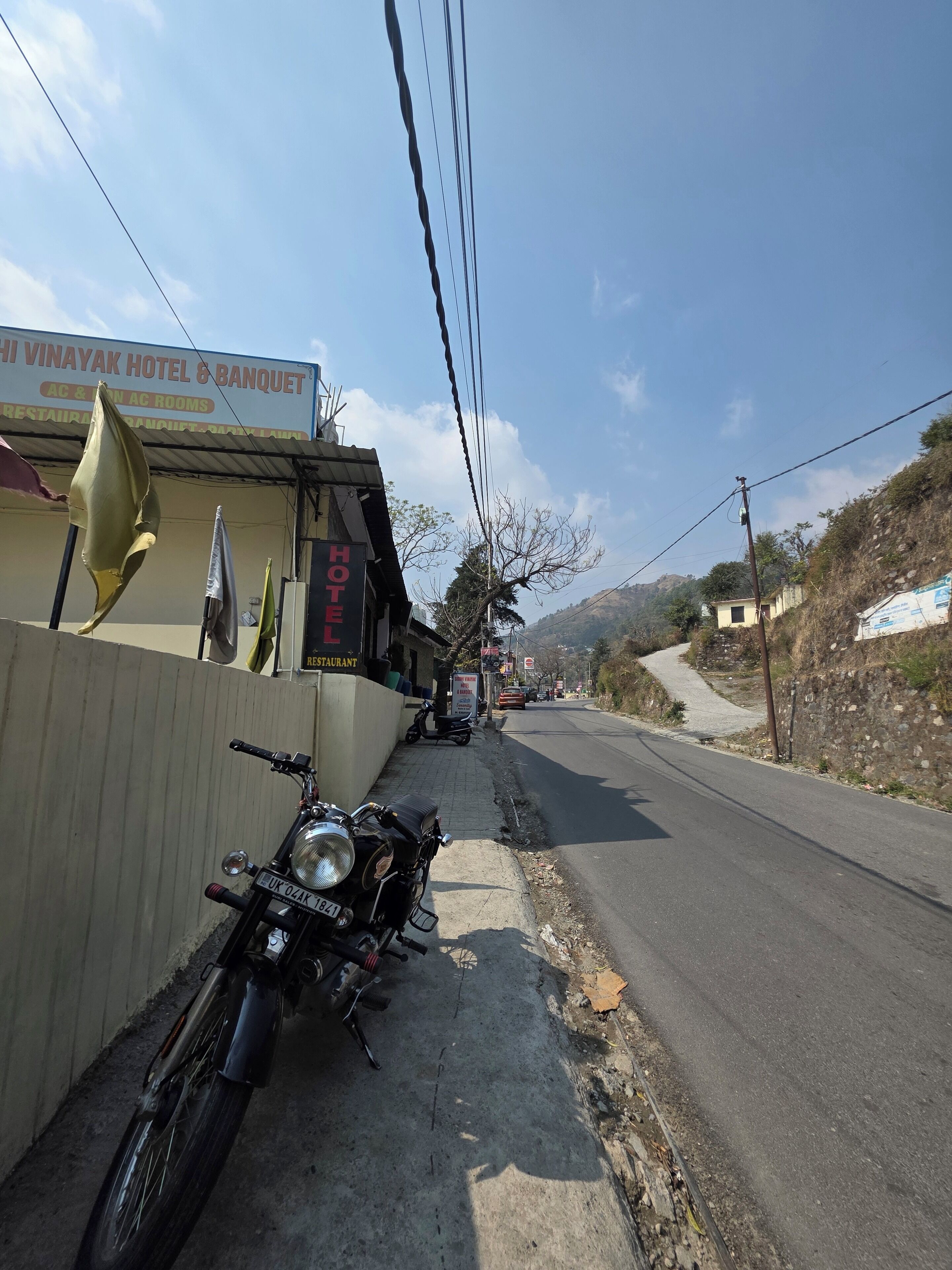 A motorcycle parked on the roadside, a sign for the HHI VINAYAK HOTEL & BANQUET, and a road leading into the distance.
