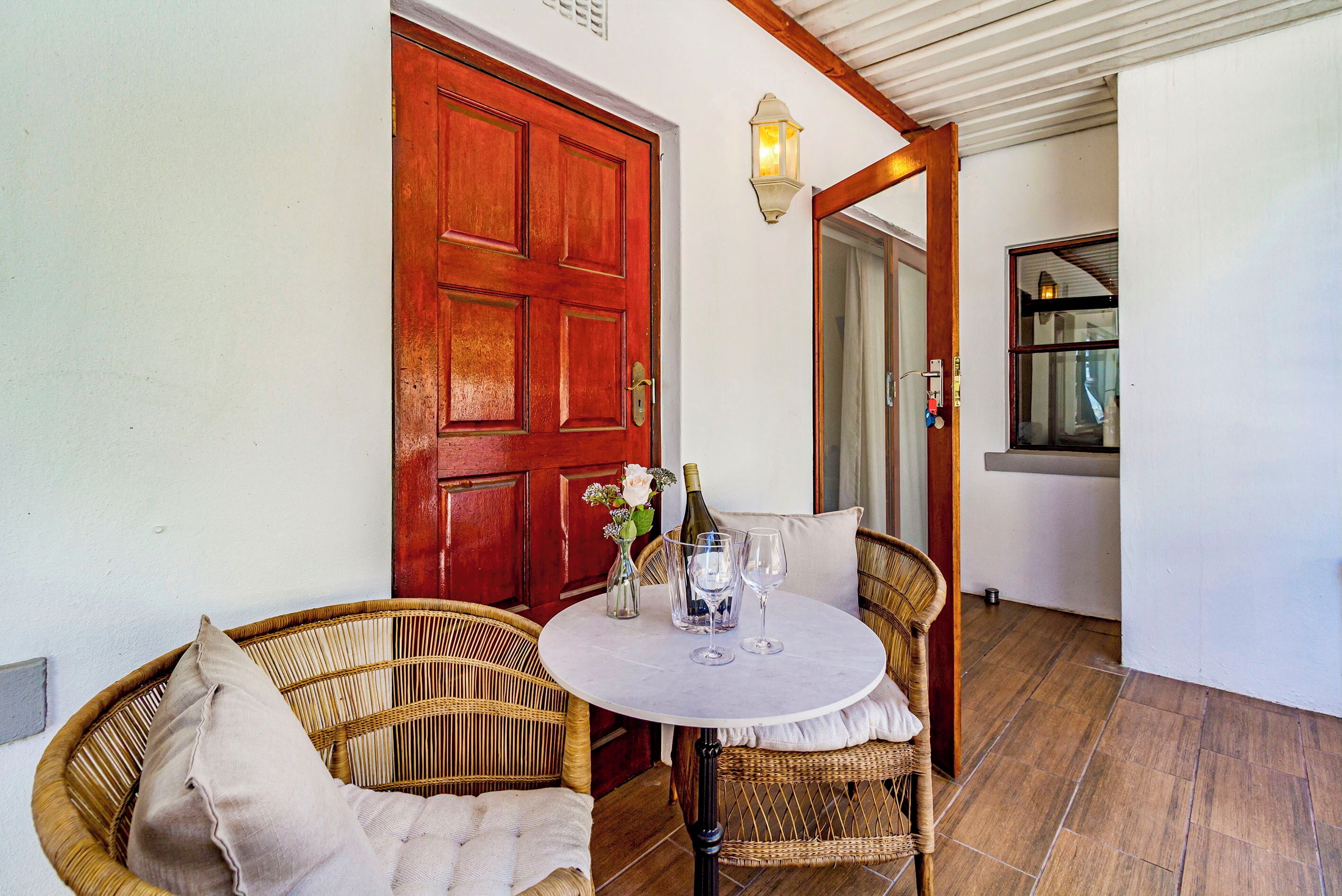 A wicker chair and table set up outside a building with a red door, a bottle, and glasses on the table.