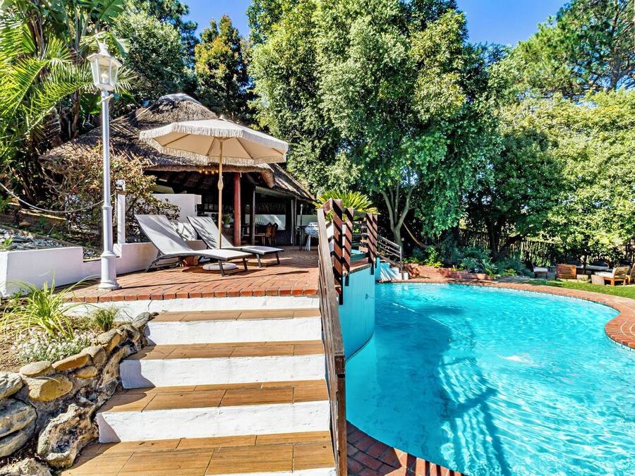A swimming pool with a wooden deck and lounge chairs, surrounded by lush greenery and a thatched-roof structure.