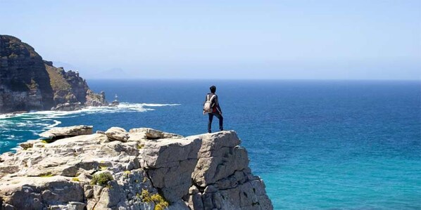 A person standing on a rocky outcrop overlooking a coastal area with clear blue water and distant land. - Villa Helderberg (Cape Town)