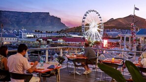 A restaurant deck with tables and chairs, overlooking a cityscape with a prominent Ferris wheel and mountains in the background. - Villa Helderberg (Cape Town)
