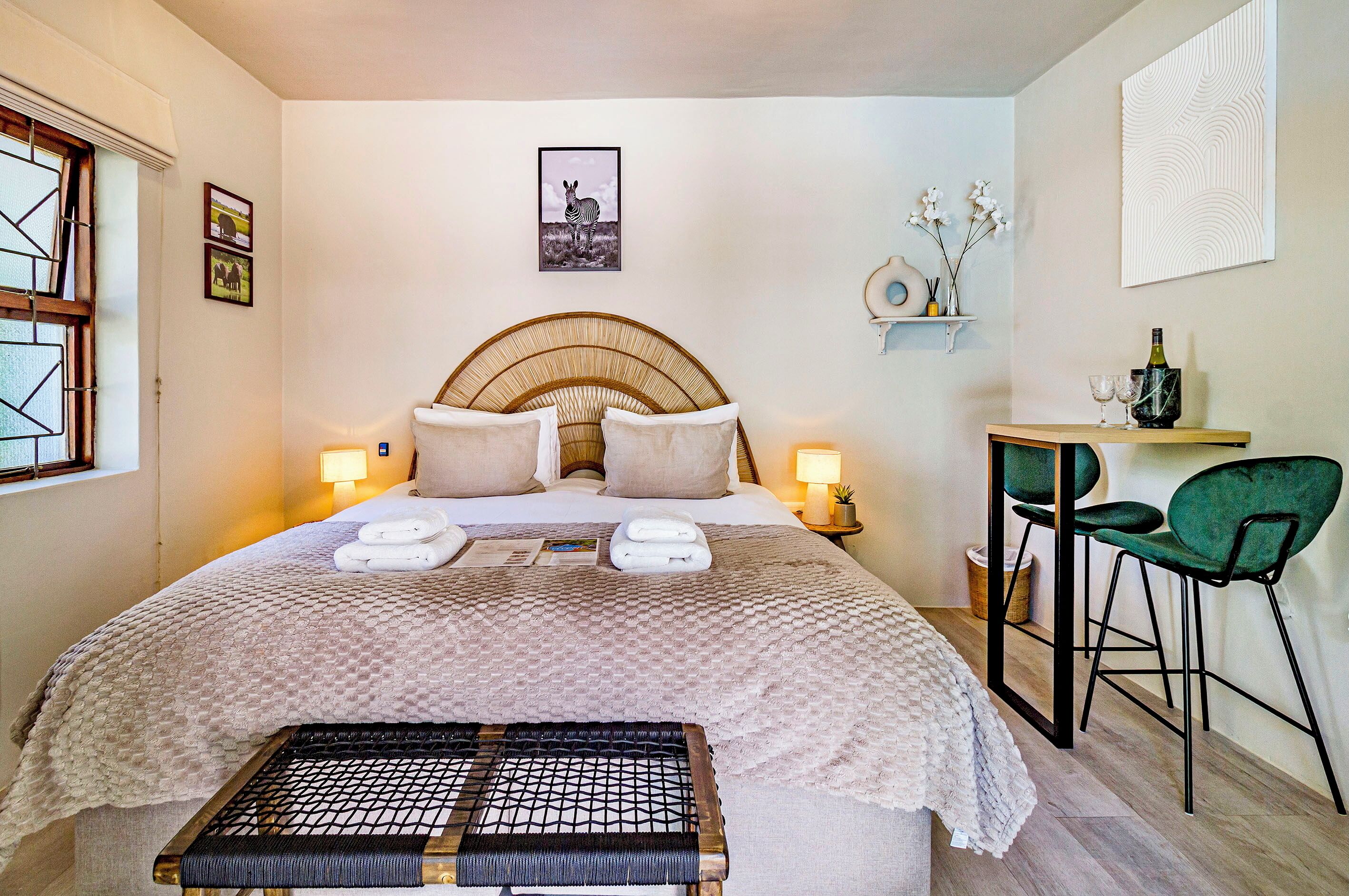 A neatly arranged bedroom with a bed, bedside lamps, a small table with chairs, and a wall-mounted shelf with decorative items.