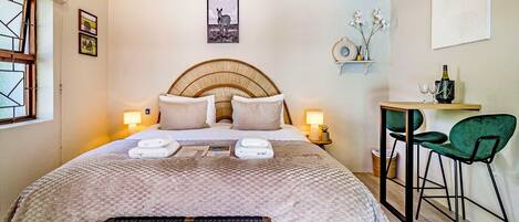 A neatly arranged bedroom with a bed, bedside lamps, a small table with chairs, and a wall-mounted shelf with decorative items.