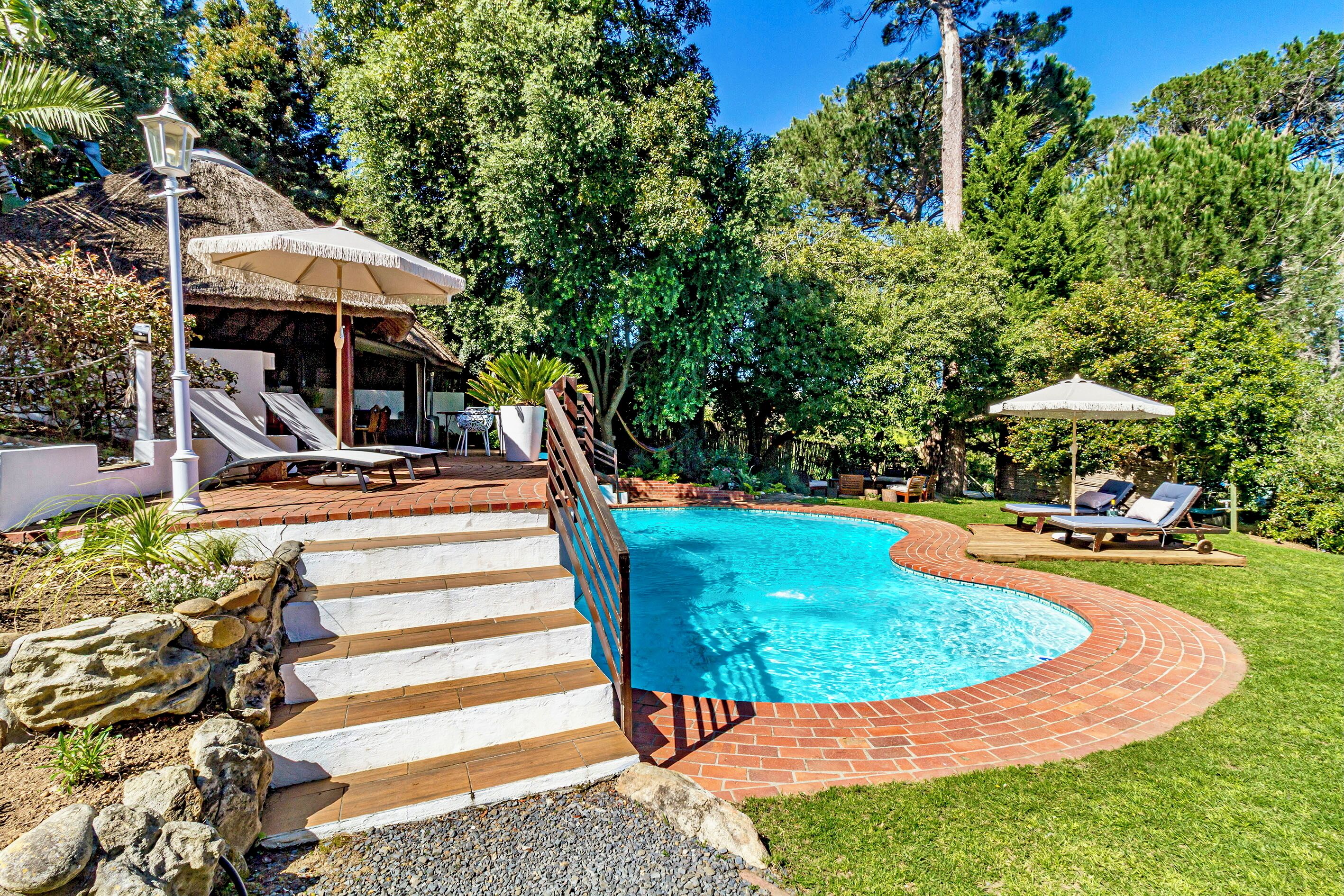 Piscina com borda curva, rodeada por um deck de tijolos e uma área gramada. Há espreguiçadeiras e guarda-sóis, e uma escadaria que leva a uma área coberta.