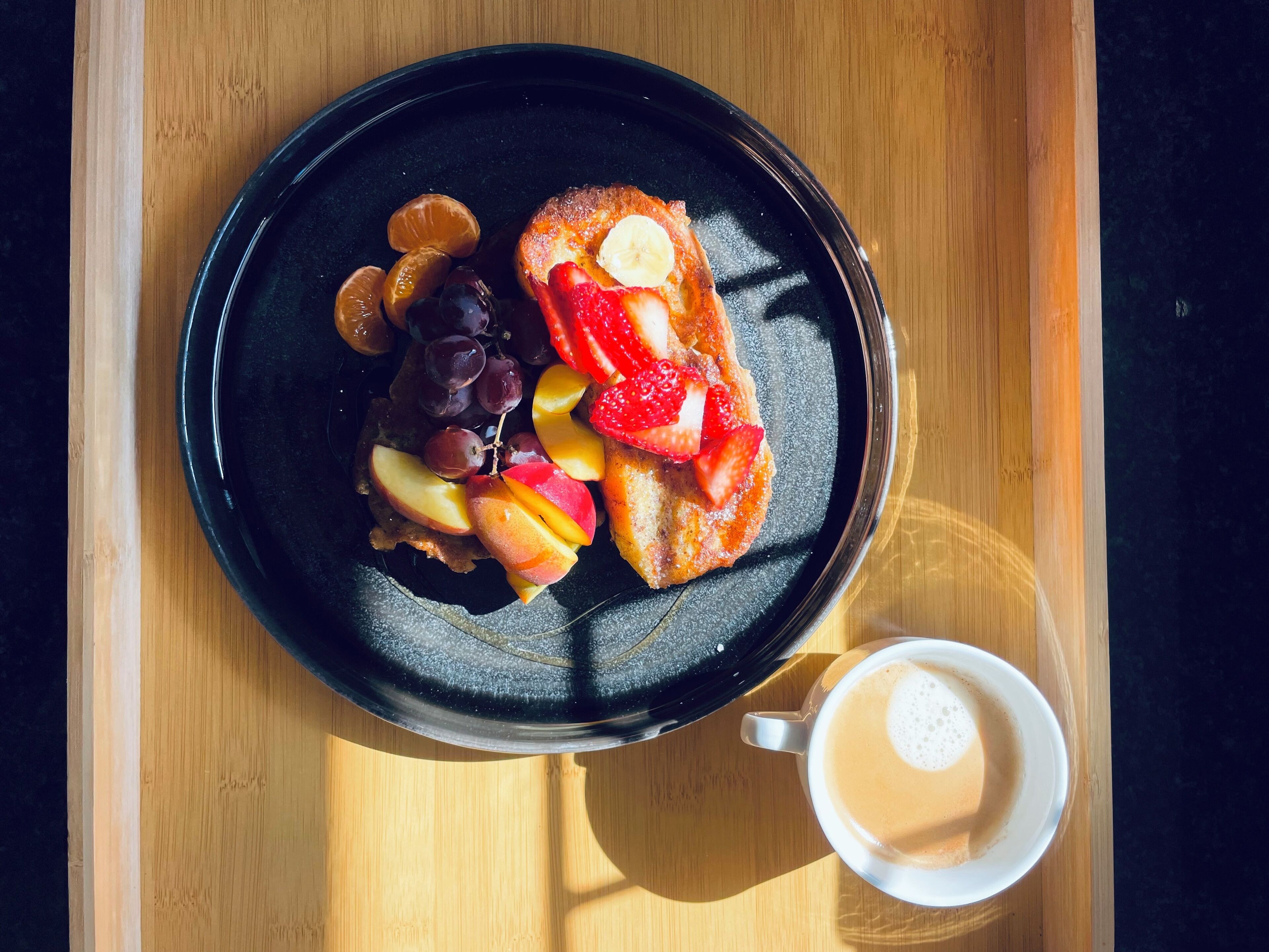 Un plato negro con tostadas cubiertas de fresas y melocotones, un tazón pequeño de uvas y una taza de café sobre una mesa de madera.