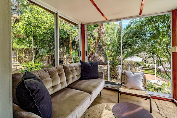 A modern living room with a beige sofa, a small wooden table, and a view of a lush garden through large glass doors. - Villa Helderberg (Cape Town)