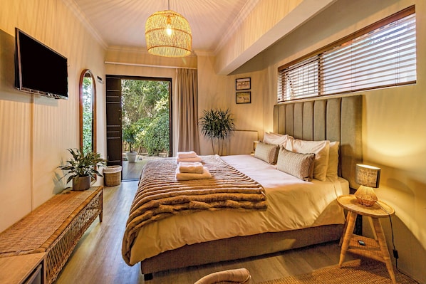 A bedroom with a large bed, a bedside table, a TV mounted on the wall, and a window with blinds.