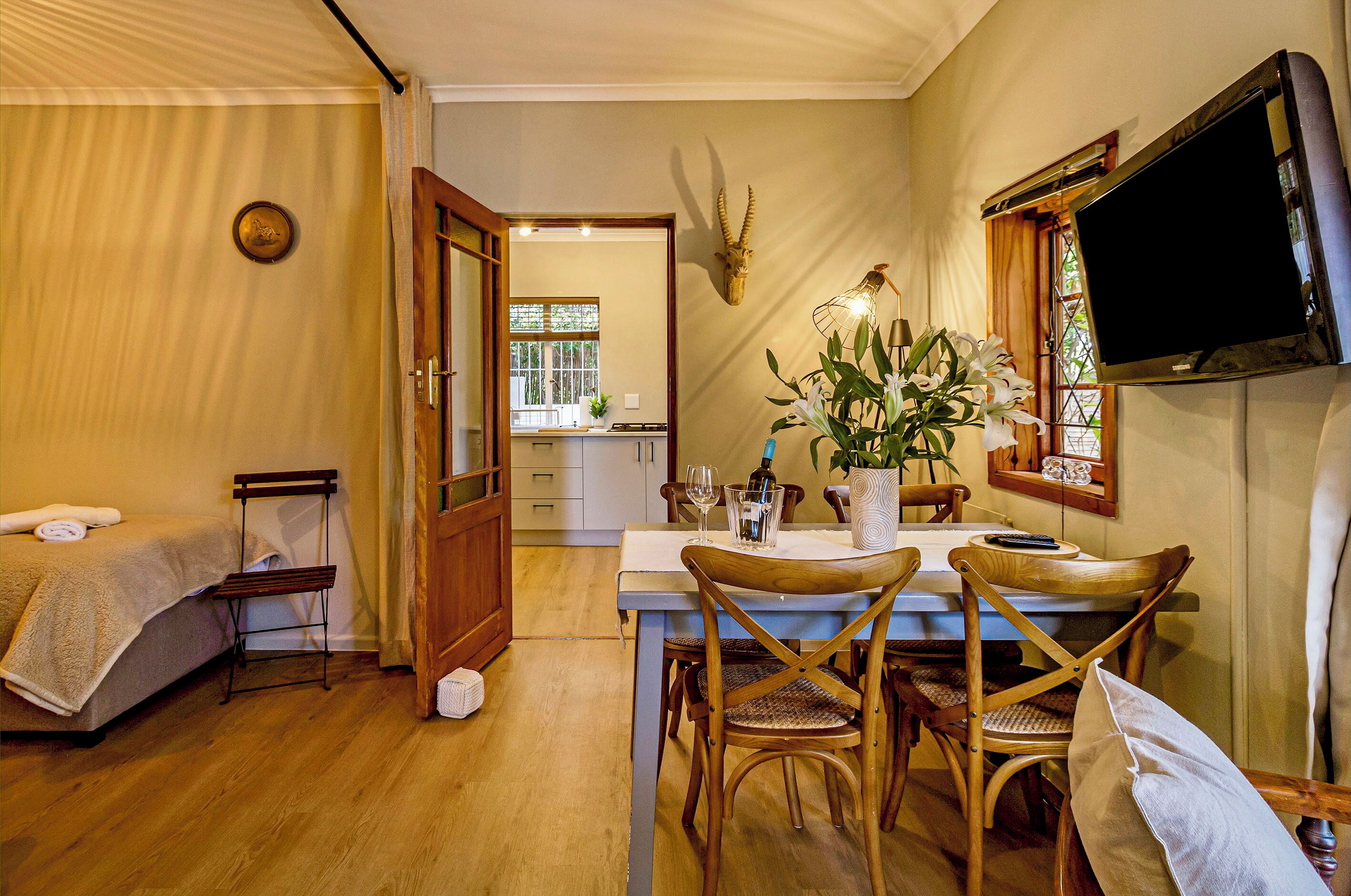 Una habitación con cama, una mesa de comedor con sillas, un televisor montado en la pared y una cocina a la vista a través de una puerta abierta.