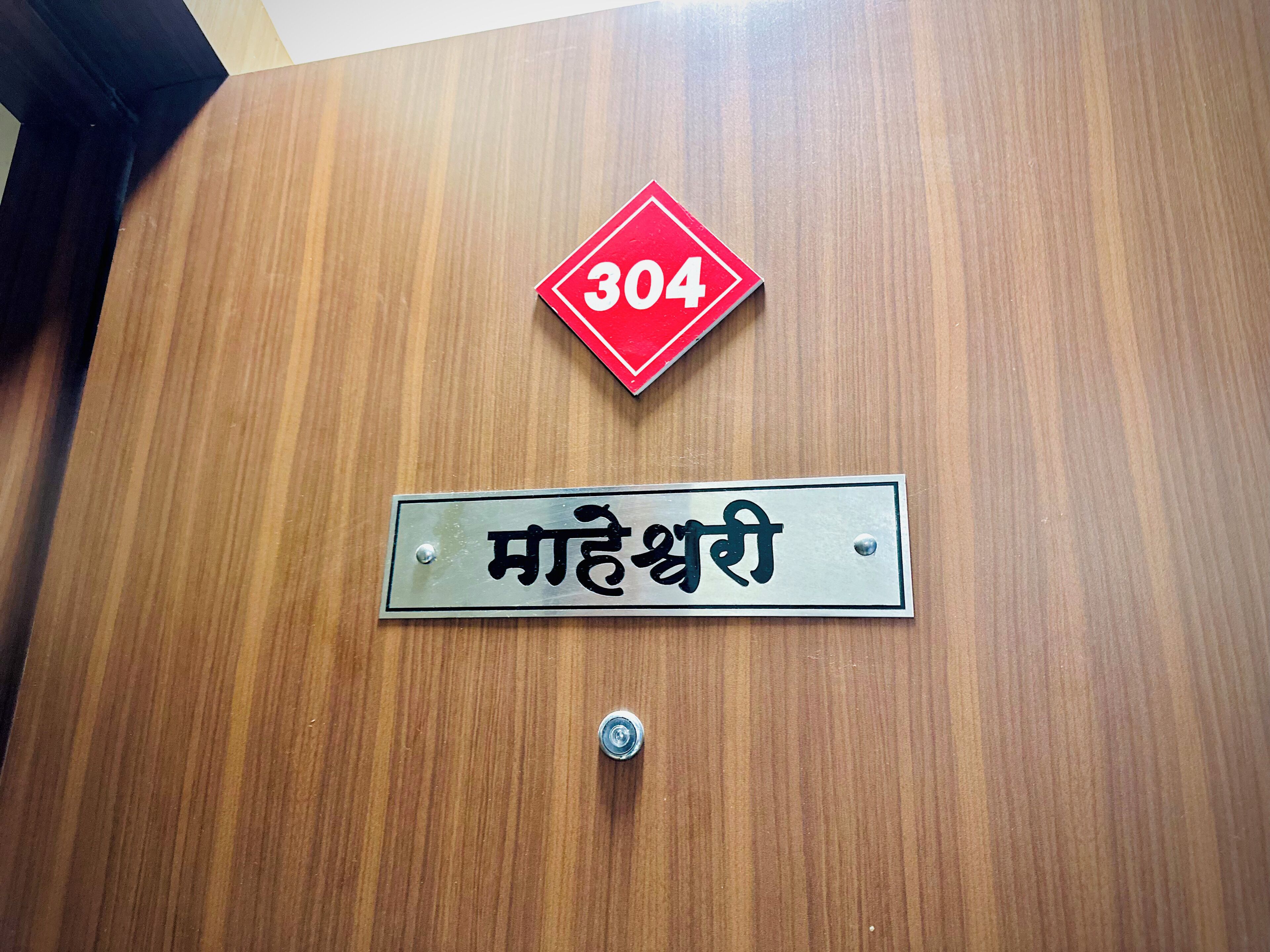 A wooden door with a red diamond-shaped sign displaying the number 304 and a metal plate with the text "महेन्द्रश्री".