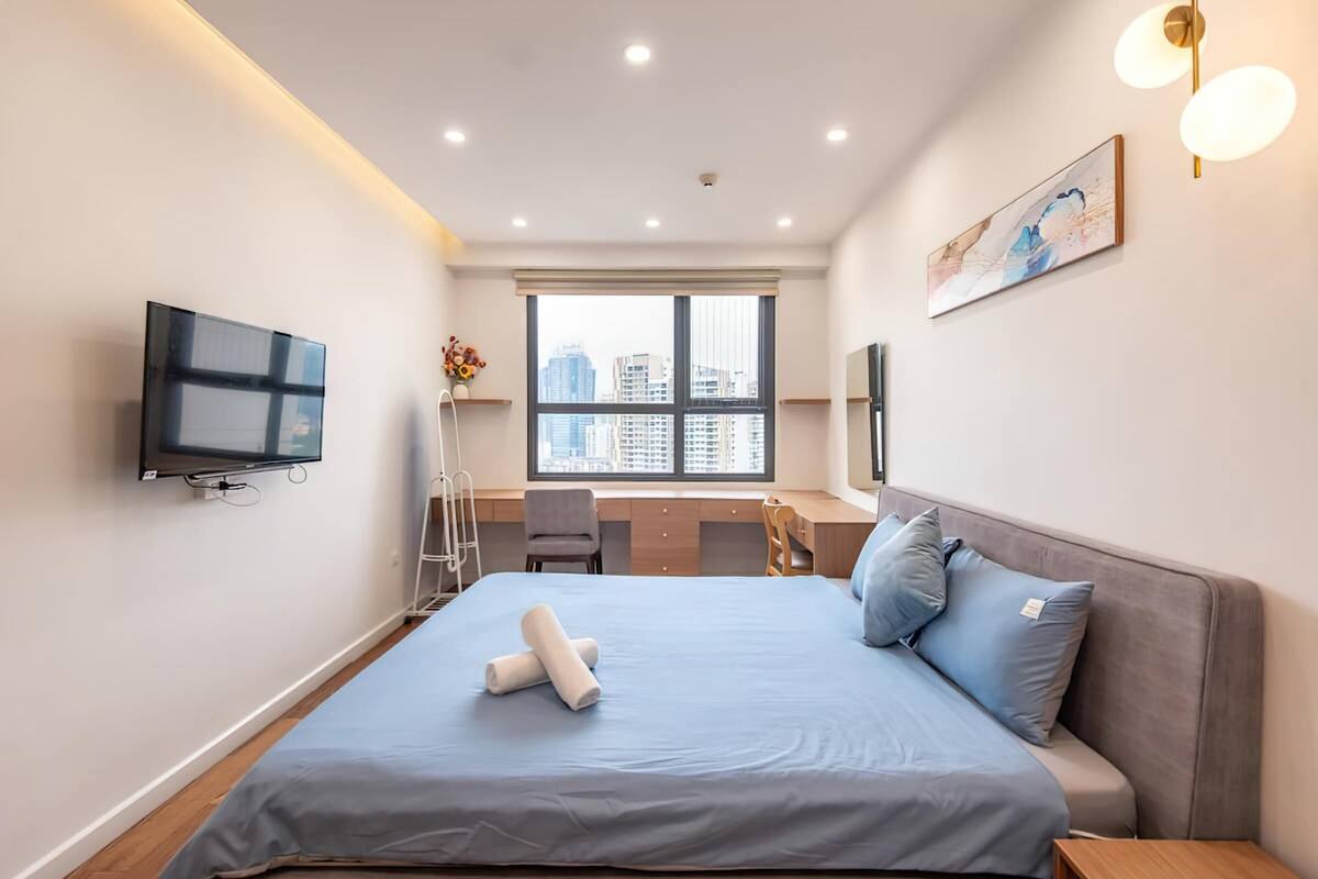 A modern hotel room with a large bed, a desk, and a view of the cityscape through the window.