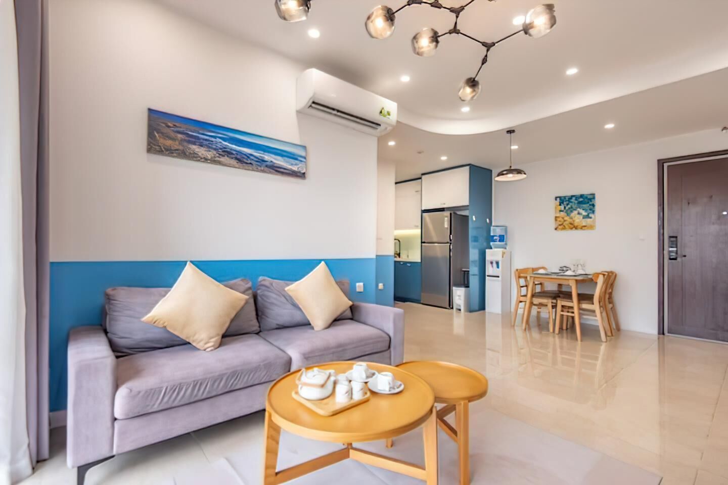 A modern living room with a sofa, a small table with towels, a dining area with a table and chairs, and a kitchen in the background.