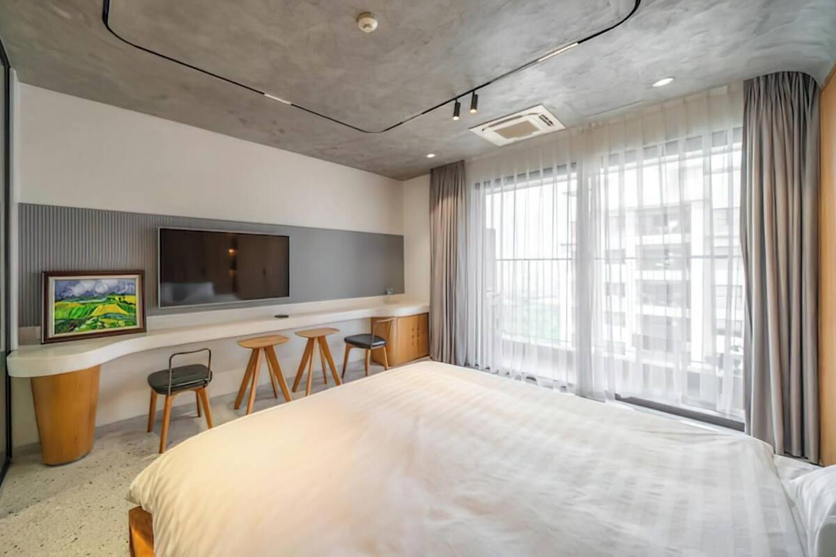 A modern hotel room with a large bed, a flat-screen TV mounted on the wall, a small desk with stools, and large windows with sheer curtains.