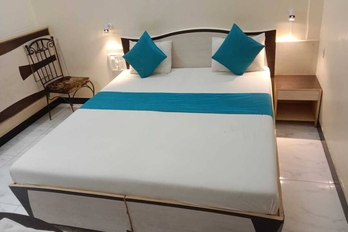 Una cama con colchón blanco, colcha azul y dos almohadas azules. A la derecha de la cama hay una mesita de noche de madera.