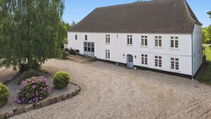 Ein weißes Gebäude mit einem Ziegeldach, einer Schotterauffahrt und einem Garten mit Sträuchern und Blumen.