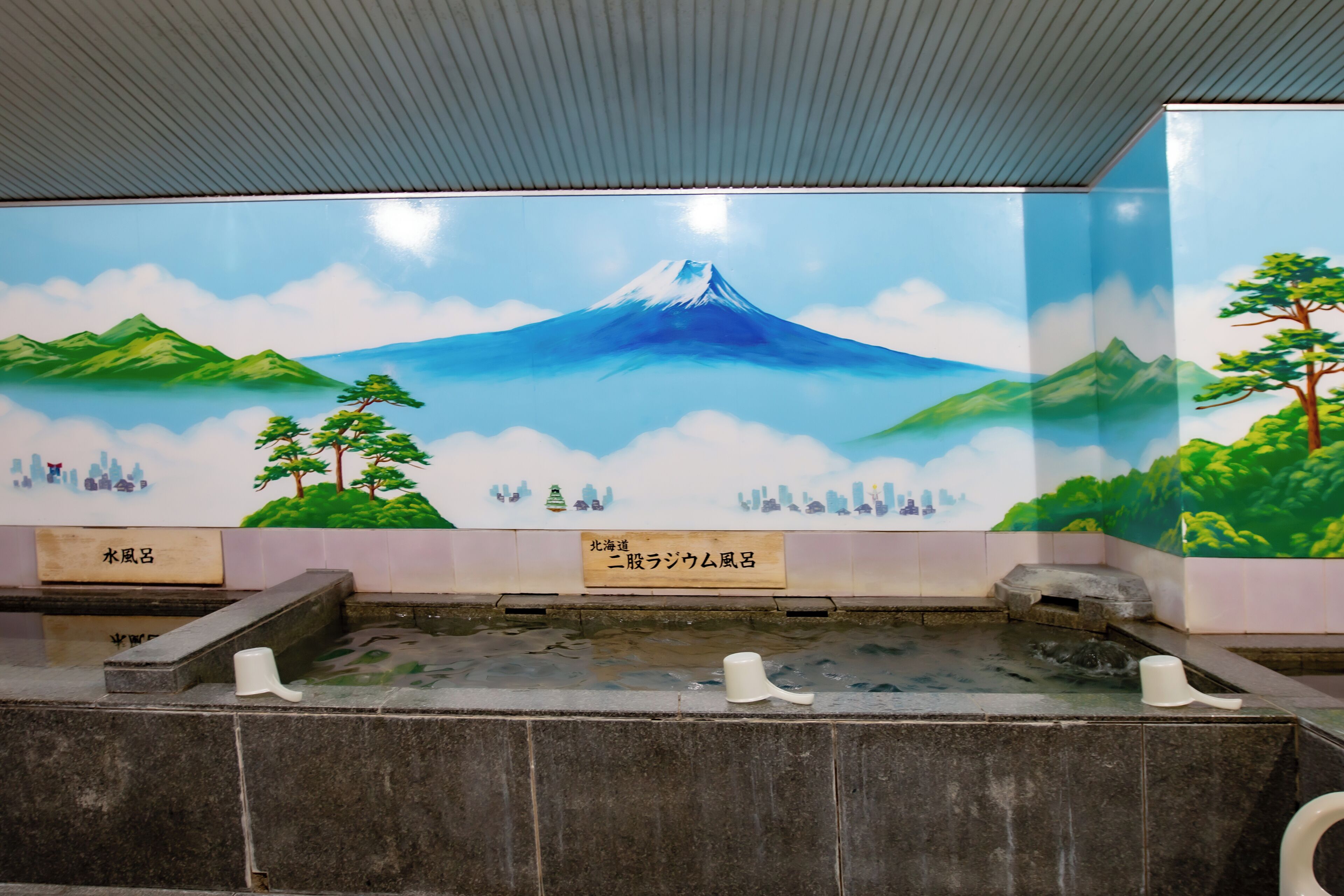 Un área de aguas termales con un mural del Monte Fuji, bancos de piedra y suelo de baldosas.