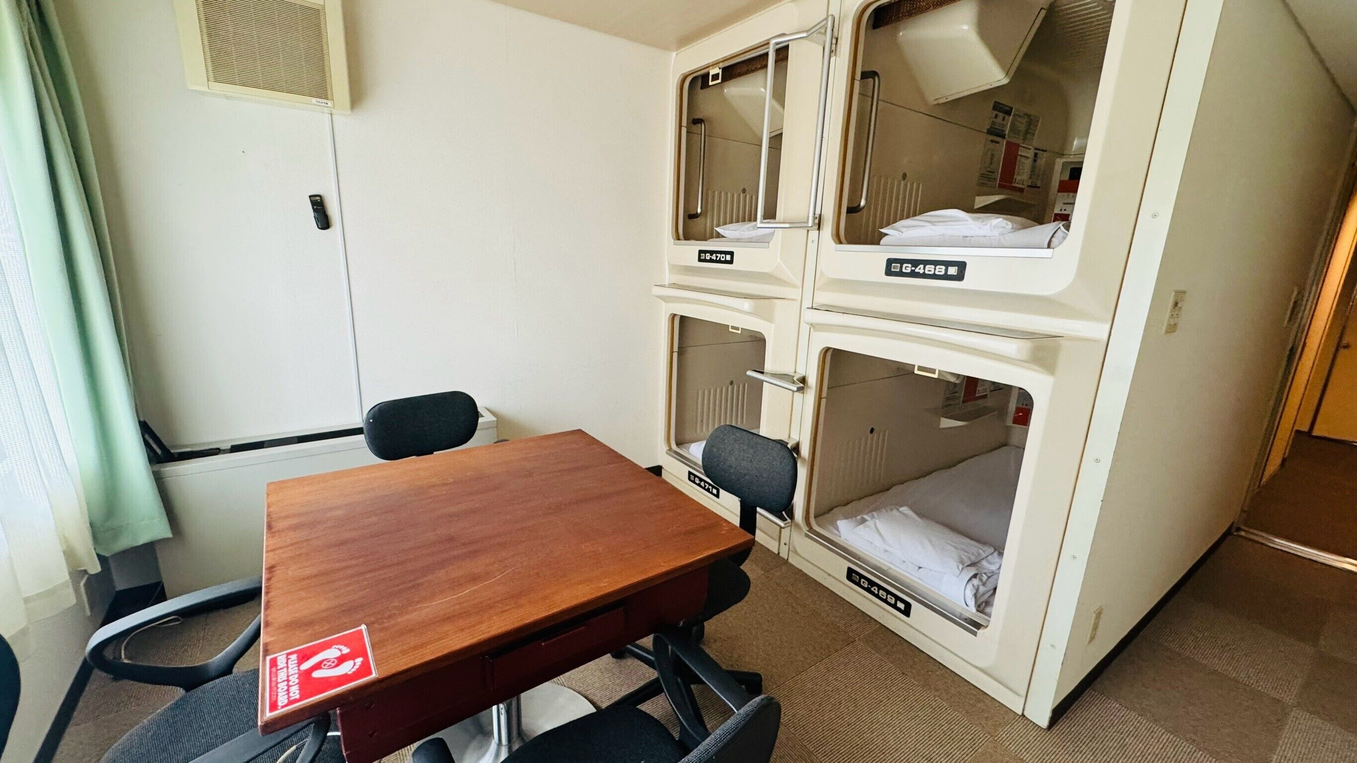 A compact hotel room with a wooden desk, two black office chairs, and a bunk bed with a small window.