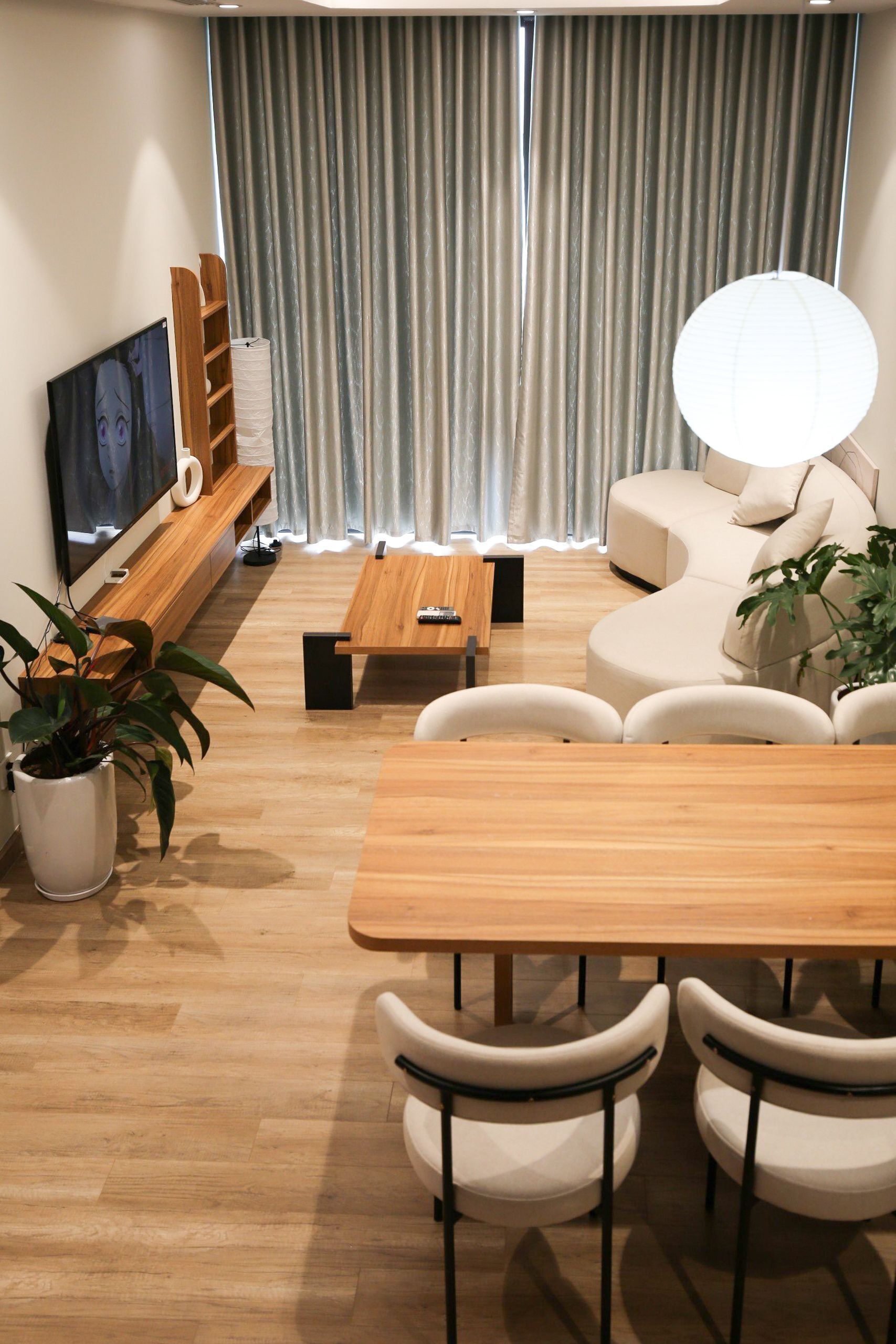 En moderne stue med et træbord, hvide stole, en beige sofa, et fjernsyn på et træstel og et stort vindue med gardiner.