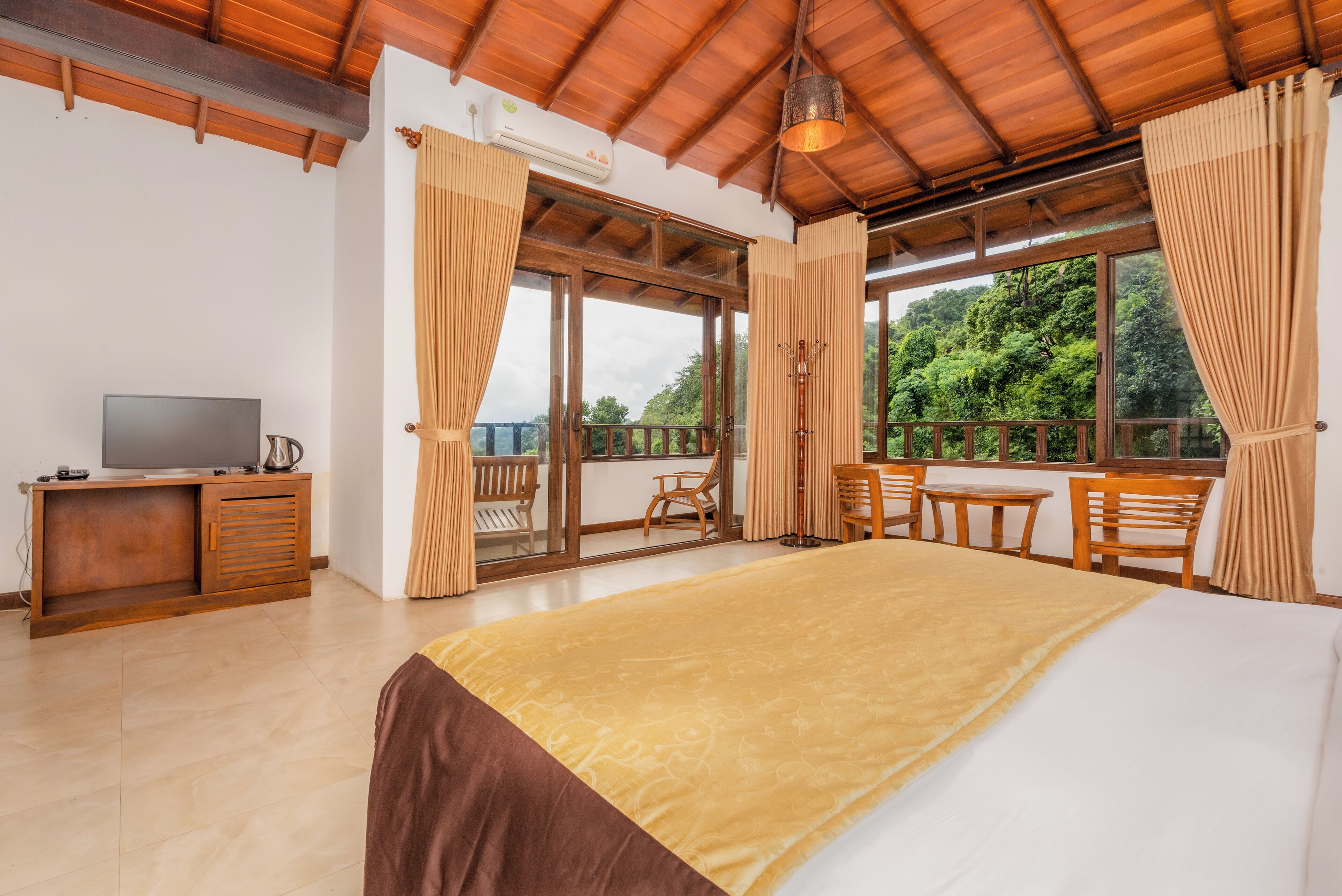 A bedroom with a large bed, wooden furniture, a TV, and a balcony with a view of greenery.