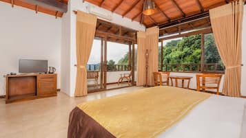 A bedroom with a large bed, wooden furniture, a TV, and a balcony with a view of greenery.