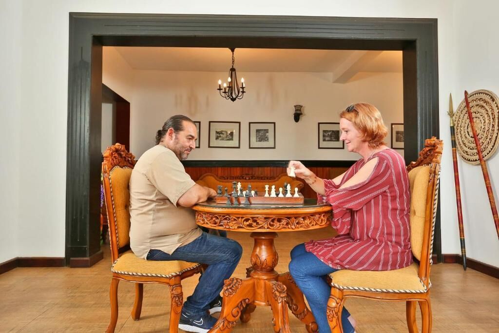 Zwei Personen spielen Schach an einem Holztisch mit Stühlen, in einem Raum mit gerahmten Bildern an der Wand und einer Kandelaber.