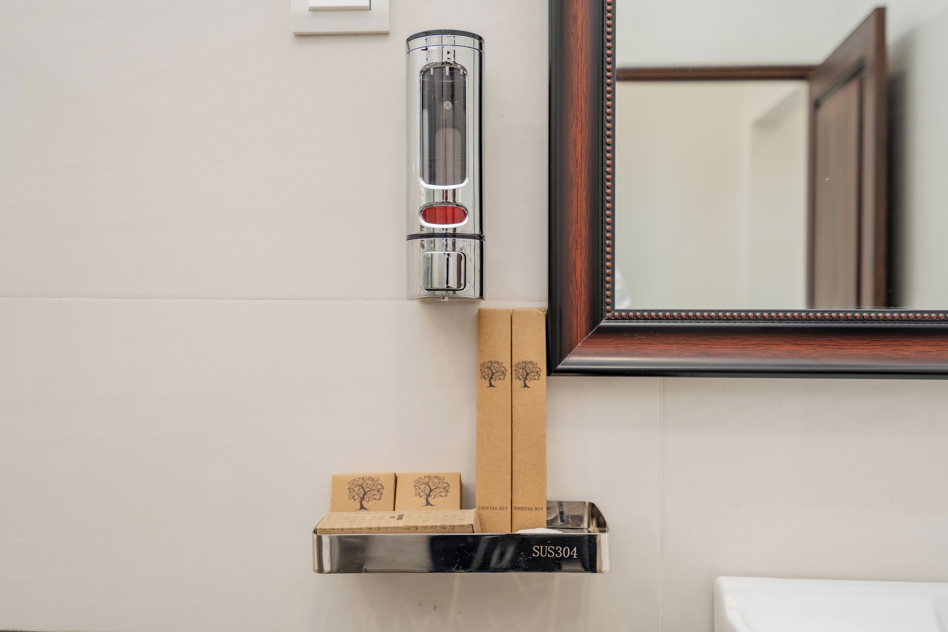 Un bagno con un distributore a parete, uno specchio e uno scaffale con porta spazzolini e una scatola con l'etichetta 'SUS304'.