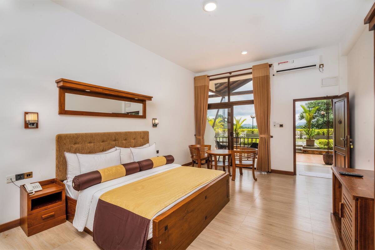 Une chambre avec un grand lit, une table de chevet en bois, une commode, un miroir et une vue sur l’extérieur grâce à une grande fenêtre.