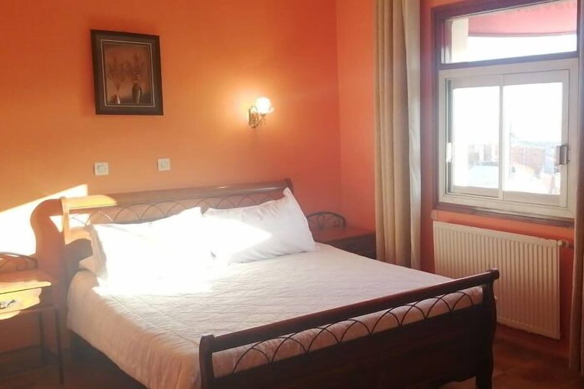 Ein Schlafzimmer mit Bett, Nachttisch, Wandlampe, Fenster mit Vorhängen und Heizkörper.