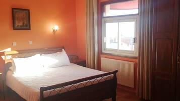 A bedroom with a bed, a nightstand, a wall-mounted lamp, a window with curtains, and a radiator.