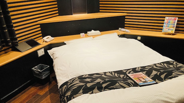 Een hotelkamer met een houten hoofdeinde, een bed met witte lakens, een tijdschrift op het bed, een telefoon op het nachtkastje en een prullenbak.