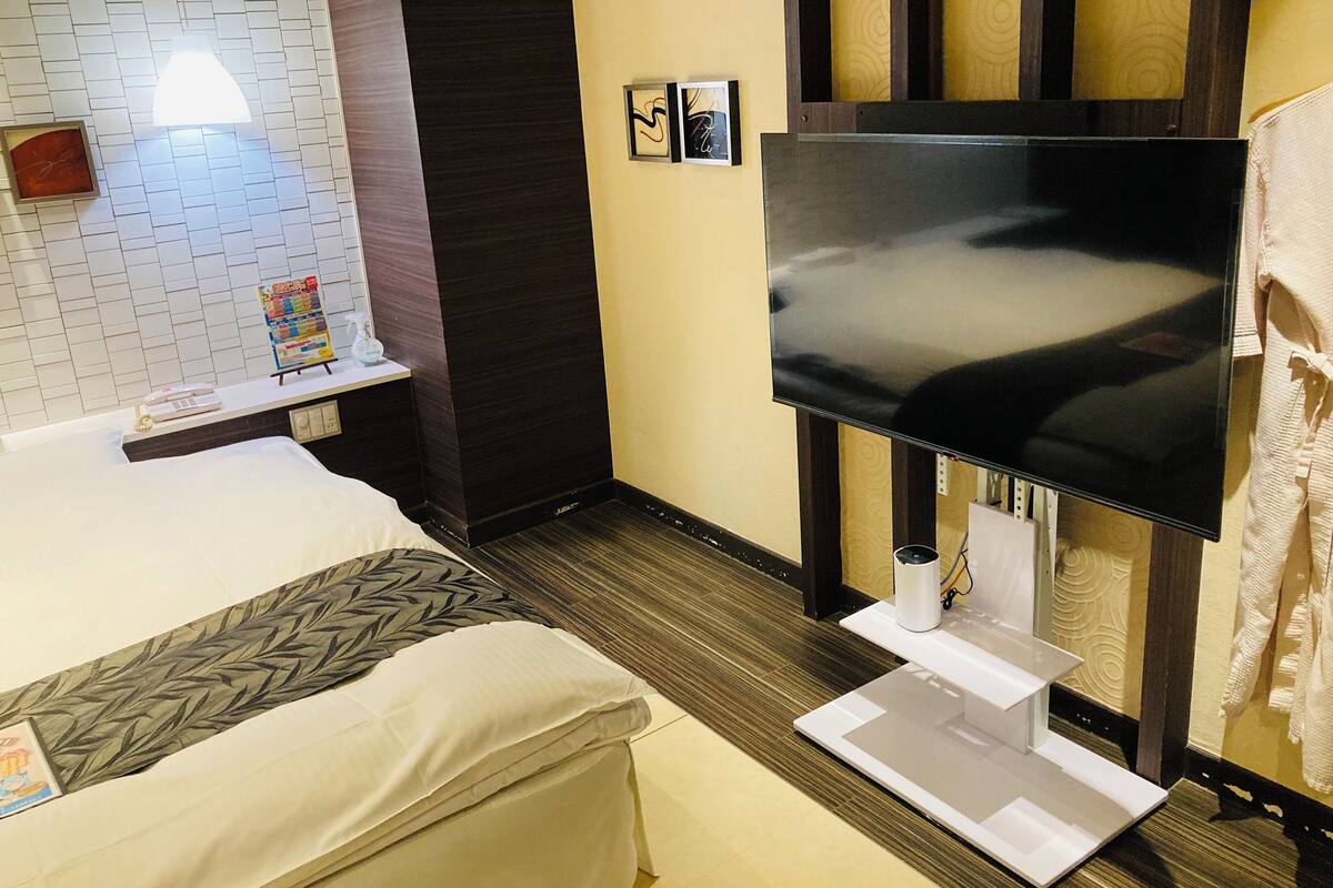 Quarto de hotel com cama, televisão de tela plana na parede, pequena mesa com controle remoto e prateleira com revista e borrifador.