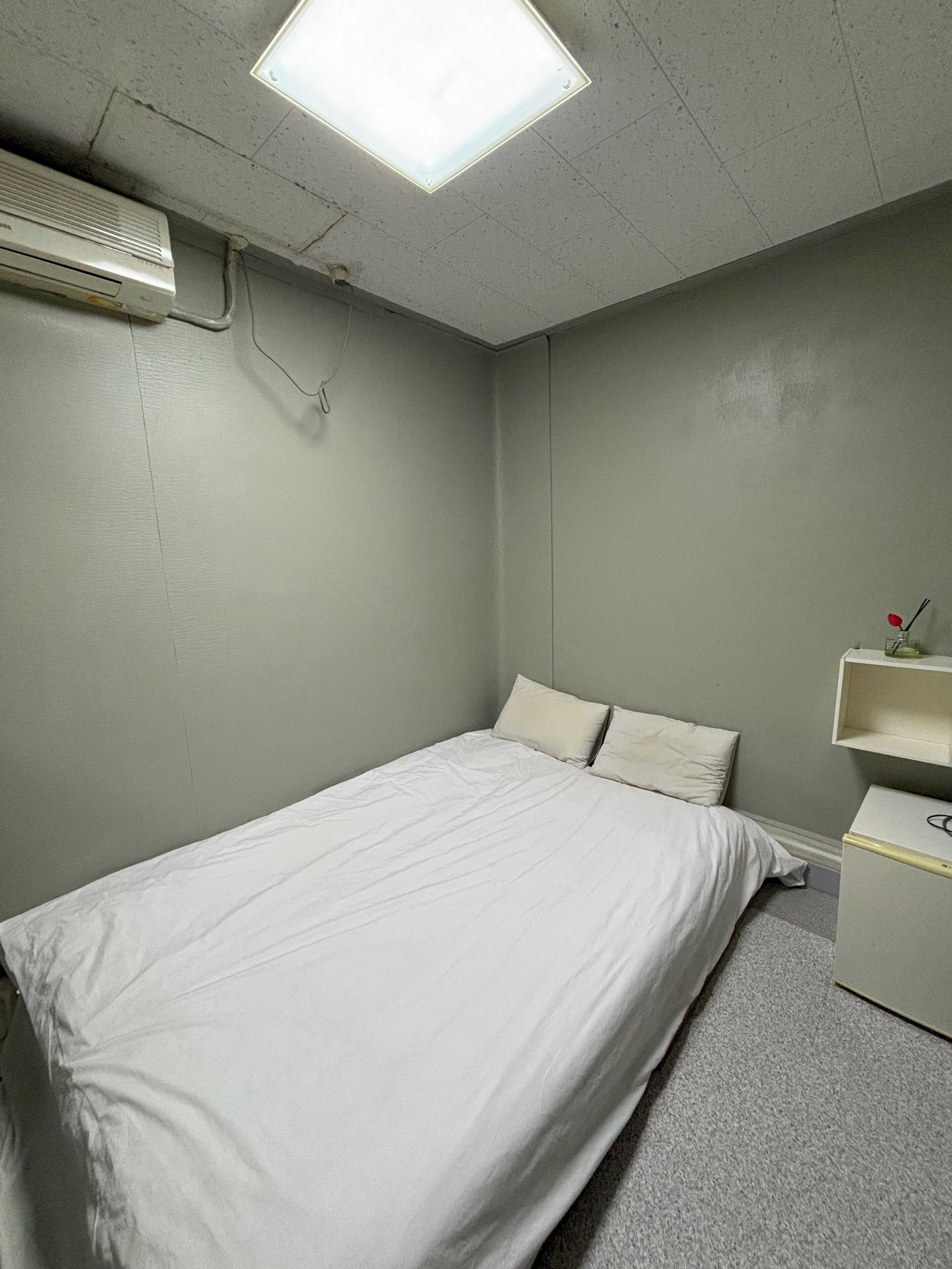 A small, sparsely furnished room with a bed, a desk, and a ceiling light.