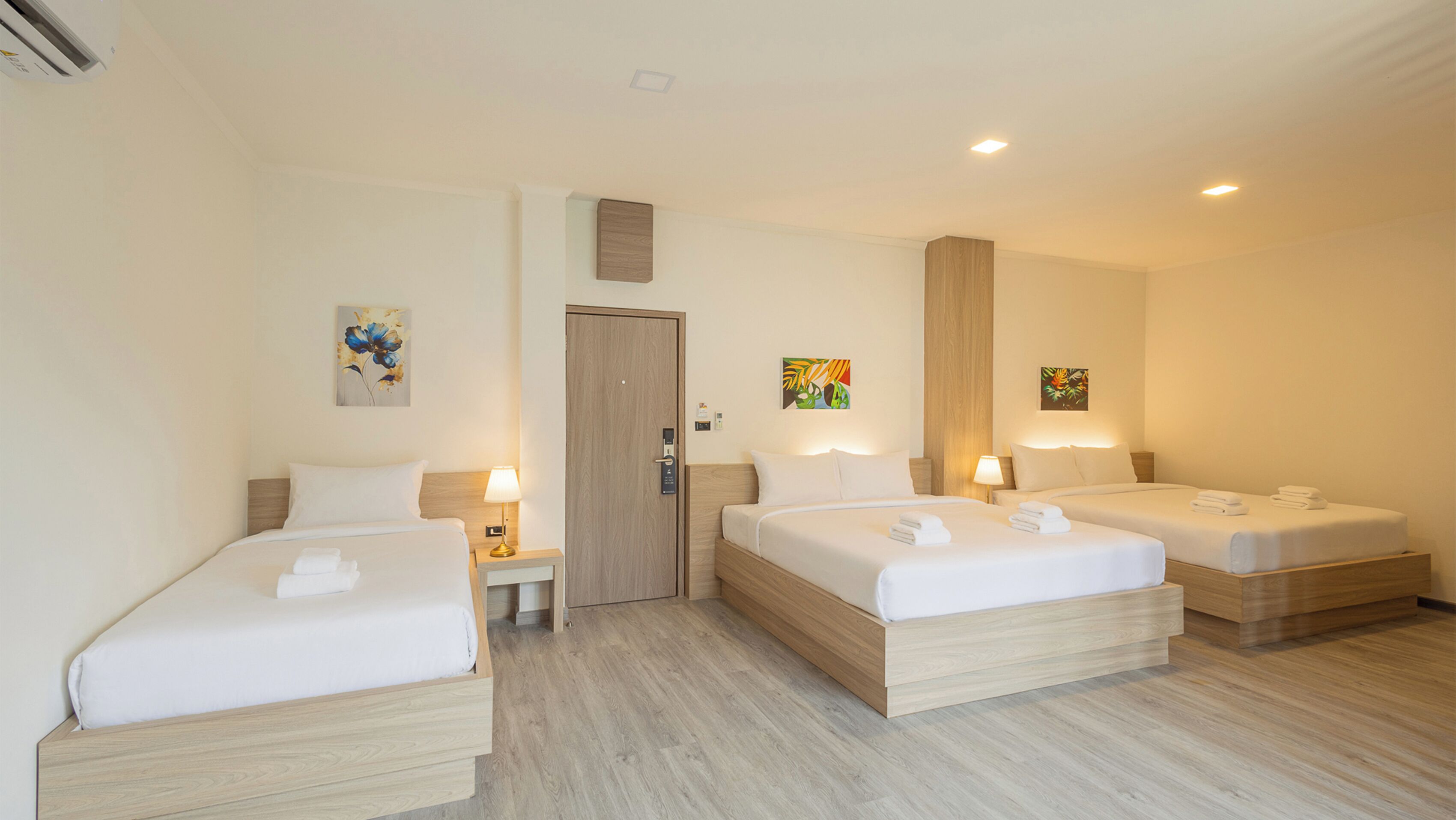 Habitación de hotel con dos camas individuales y una cama más grande, cabeceros de madera, una puerta y dos cuadros colgados en la pared.