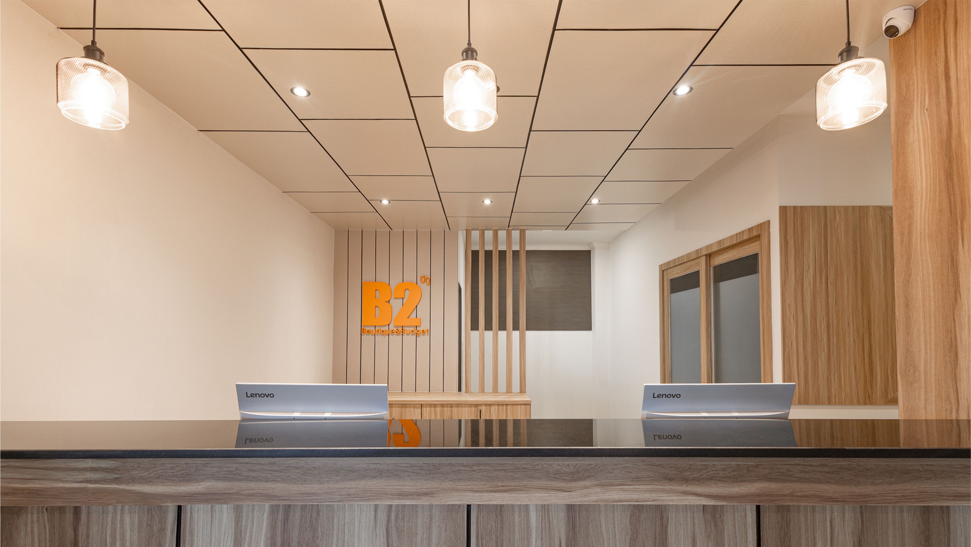 Zona de recepción con dos computadoras Lenovo sobre un mostrador, un letrero con 'B2' y 'Boutique & Budget', y un panel de pared de madera.