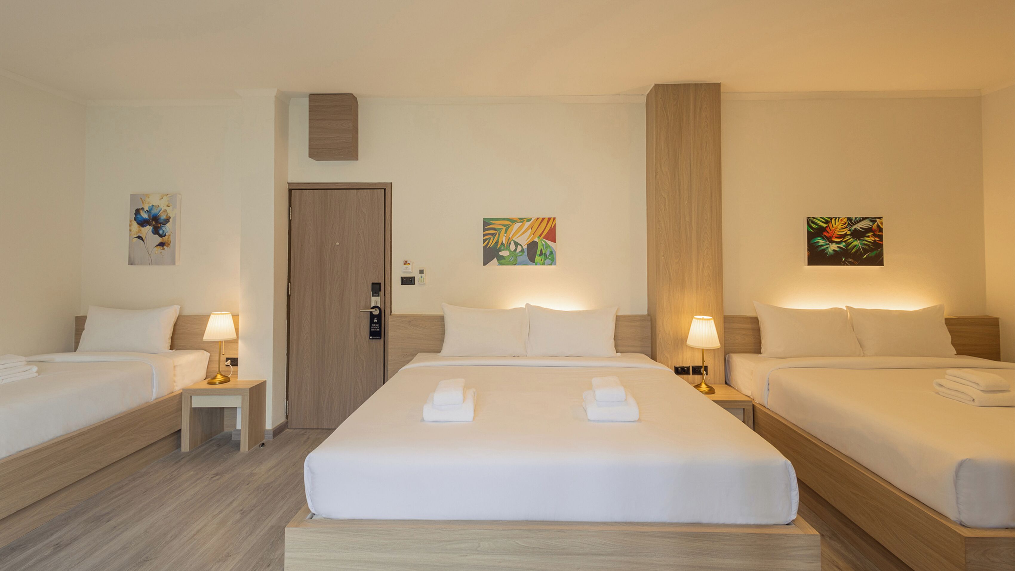 A hotel room with two single beds and a larger bed, all neatly made with white linens and towels. There are small bedside tables with lamps, and the room has a wooden floor and walls.