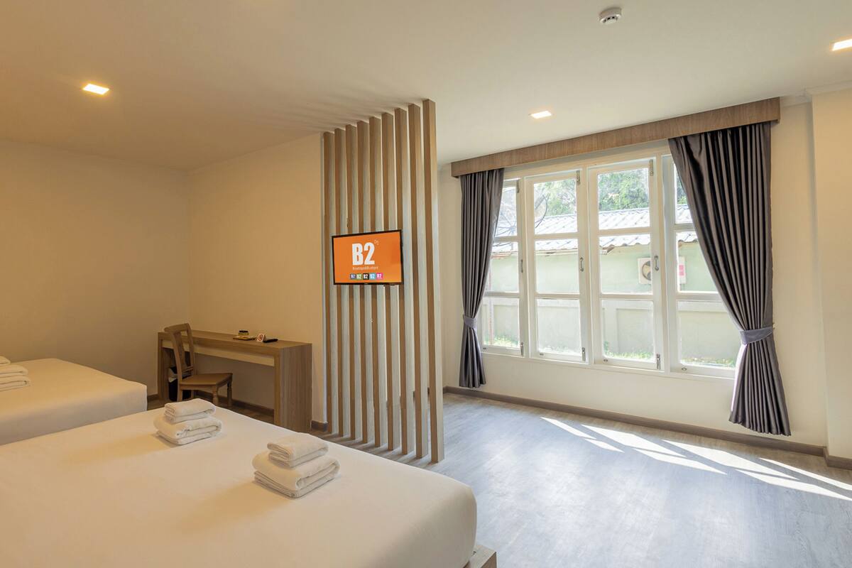 A hotel room with two beds, a desk, a TV screen displaying room number B2, and a window with curtains.