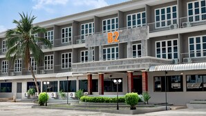 Un hotel de varios pisos con un llamativo letrero 'B2', unidades de aire acondicionado y un acceso ajardinado con palmeras y arbustos.