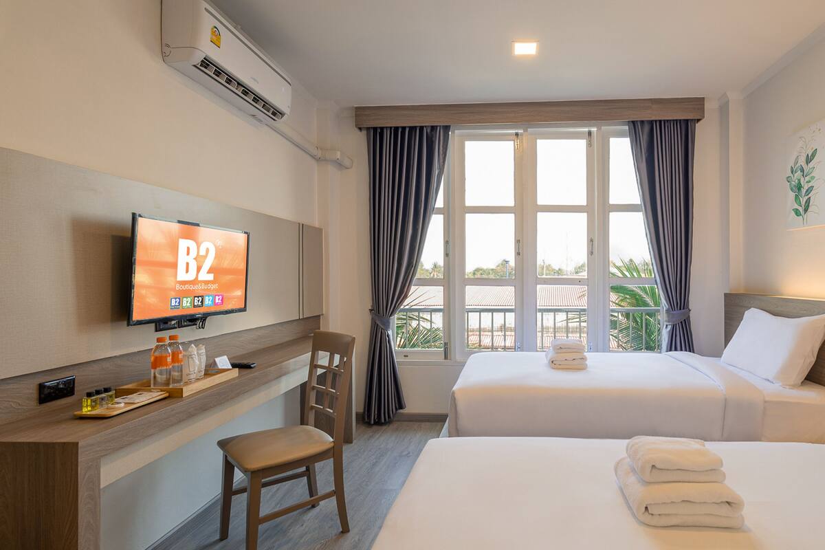 A hotel room with two beds, a desk with a TV displaying 'B2 Boutique & Budget', a chair, and a view of the outdoors through large windows.