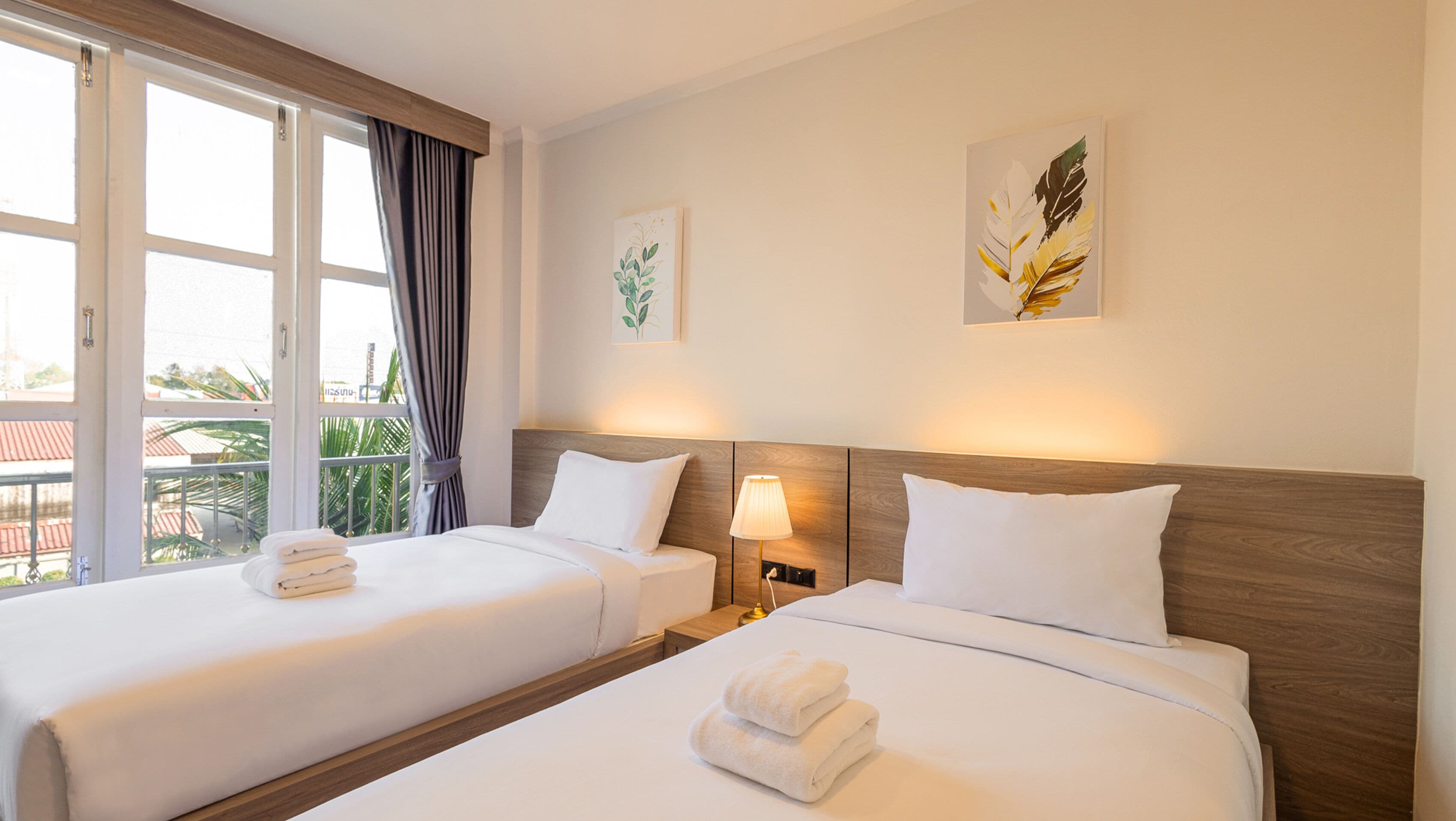 Habitación de hotel con dos camas, una ventana con cortinas, una mesita de noche con lámpara y una obra de arte en la pared.