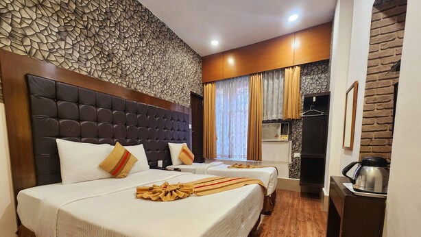 A hotel room with a large bed, a smaller bed, a wooden headboard, a stone-patterned wall, a window with curtains, and a kitchen area with a kettle. - HOTEL RAUNAK (Kolkata)