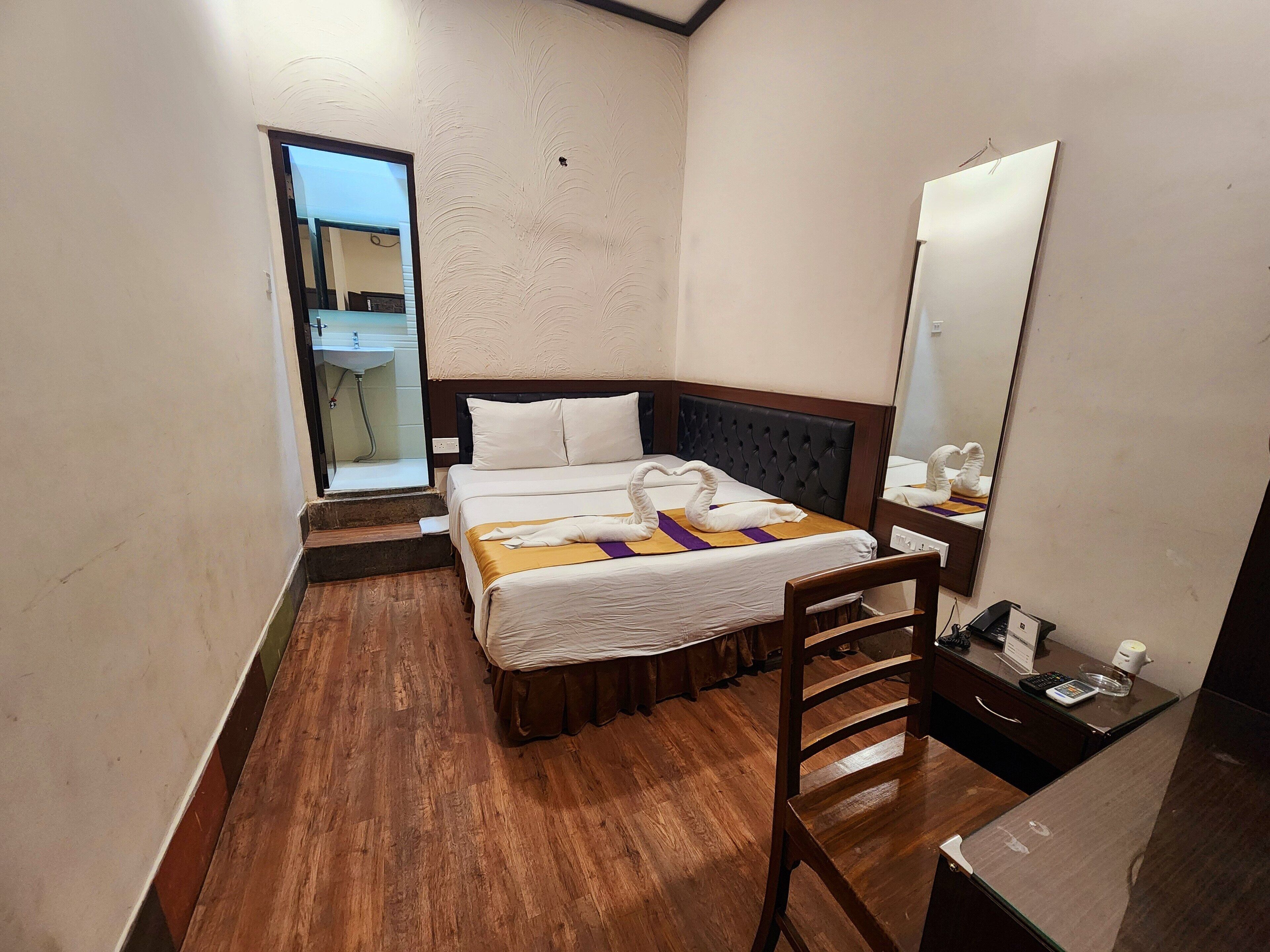 Camera d'albergo con pavimento in legno, un letto con testiera, un comodino con lampada, uno specchio e un bagno visibile attraverso una porta aperta.